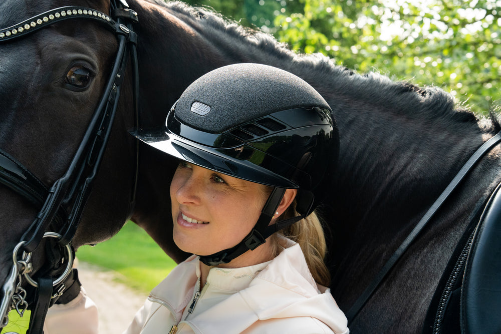 Woman wearing a black equestrian horse riding helmet standing next to a black horse in a natural setting.